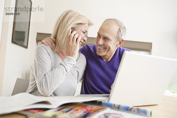 Frau,  die mit ihrem Mann auf einem Handy vor einem Laptop sitzt.