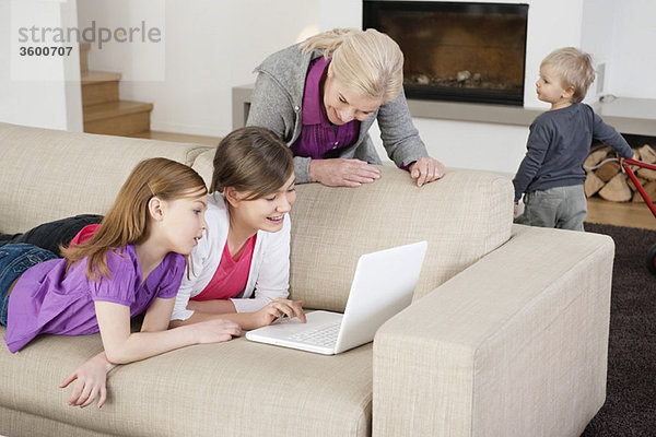 Zwei Mädchen,  die einen Laptop auf einer Couch benutzen,  während ihre Großmutter neben ihnen steht.