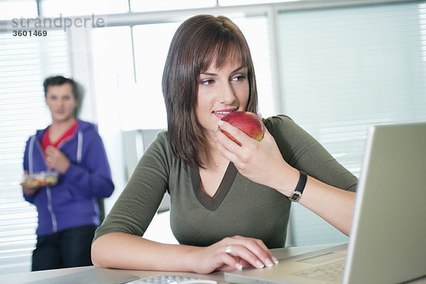 Geschäftsfrau,  die einen Apfel isst und einen Laptop benutzt