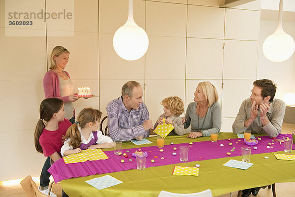 Familie bei einer Geburtstagsfeier mit einer Frau mit Kuchen im Hintergrund