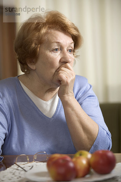 Eine besorgte ältere Frau,  die mit der Hand am Tisch sitzt und ihren Mund bedeckt.