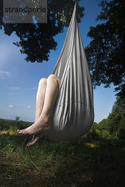 Eine Frau sitzt in einer geschlossenen Hängematte,  die an einem Baum hängt,  nur die Beine sind sichtbar.