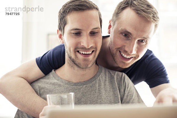 Glückliches homosexuelles Paar,  das den Laptop benutzt,  während es sich zu Hause umarmt.
