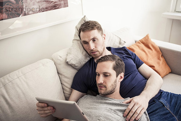 Junge Homosexuelle nutzen gemeinsam digitale Tabletten und entspannen sich zu Hause auf dem Sofa.