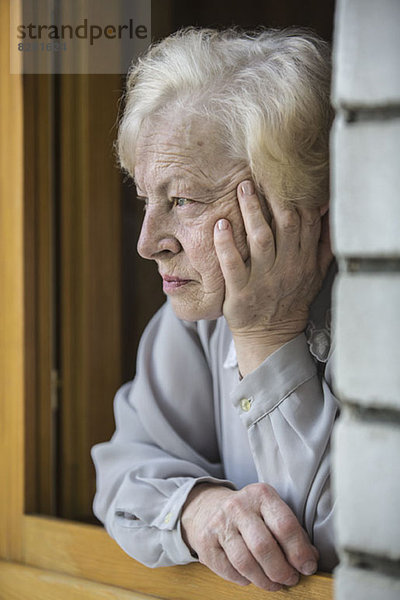 Eine ältere Frau,  die sich auf ein Fensterbrett stützt und kontemplativ aussieht.