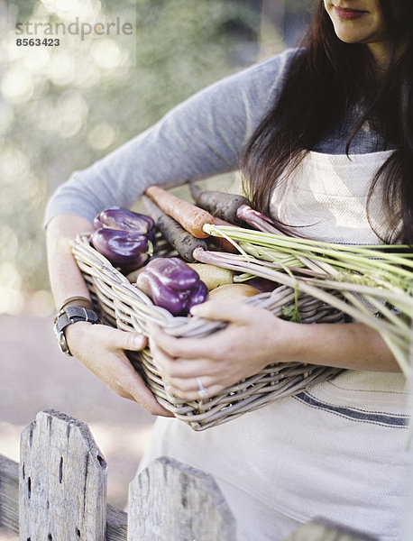 Eine junge Frau in einem Gemüsegarten,  die einen Korb mit frisch geerntetem Bio-Gemüse,  Paprika und einer Eierpflanze trägt.