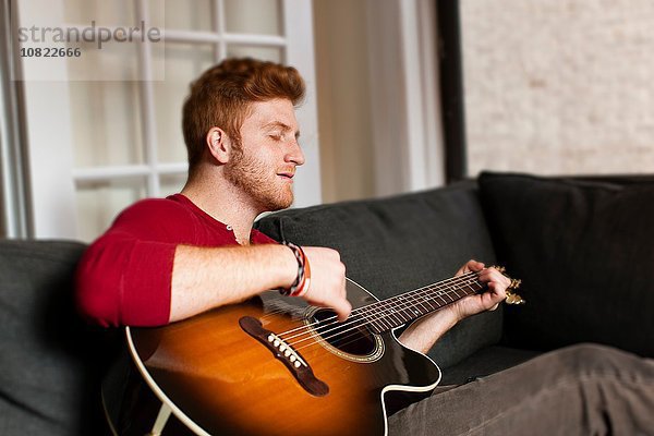 Junger Mann sitzt auf dem Sofa und spielt Gitarre,  die Augen geschlossen.