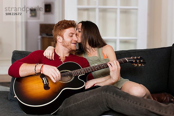 Junge Frau auf dem Sofa küsst jungen Mann,  der Gitarre auf der Wange spielt.