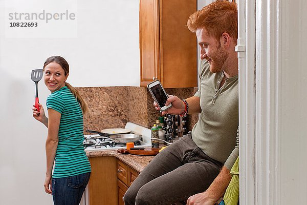 Junger Mann in der Küche mit dem Smartphone,  um die junge Frau mit dem Spachtel zu fotografieren.