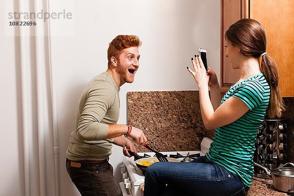Seitenansicht der jungen Frau in der Küche mit dem Smartphone,  um den jungen Mann beim Kochen zu fotografieren.