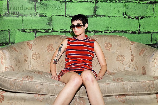 Porträt einer erwachsenen Frau mit Sonnenbrille,  die auf einem getragenen Sofa sitzt.