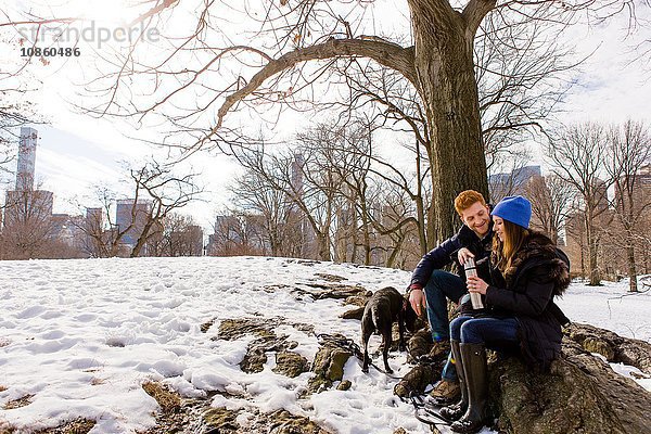 Junges Paar sitzt mit Hund und Trinkflasche im verschneiten Central Park,  New York,  USA