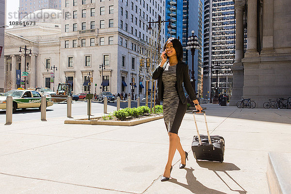 Geschäftsfrau,  die im Freien spazieren geht,  einen Koffer mit Rädern zieht und ein Smartphone benutzt