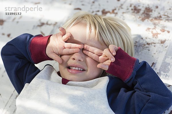 Verspielter kleiner Junge,  der sich hinlegt und seine Augen mit den Händen bedeckt.