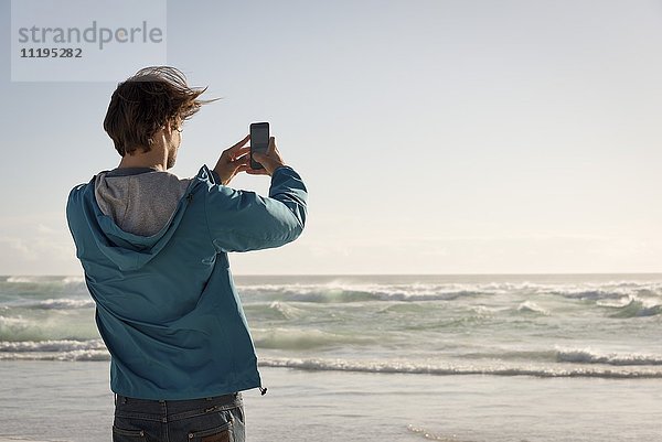 Glücklicher junger Mann beim Fotografieren mit Fotohandy am Strand