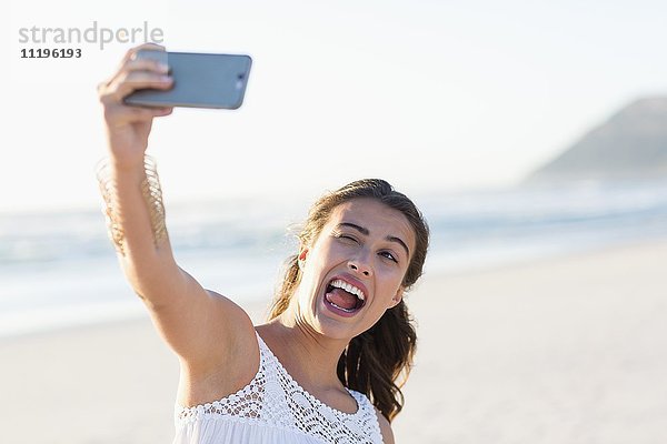 Verspielte junge Frau nimmt Selfie mit Smartphone am Strand