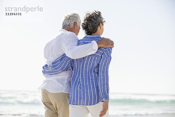 Rückansicht eines Paares,  das mit einem Arm um den anderen am Strand steht.