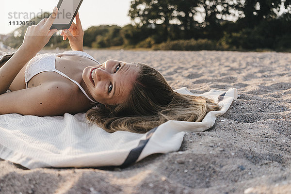 Lächelnde junge Frau auf Decke am Strand liegend mit Tablette