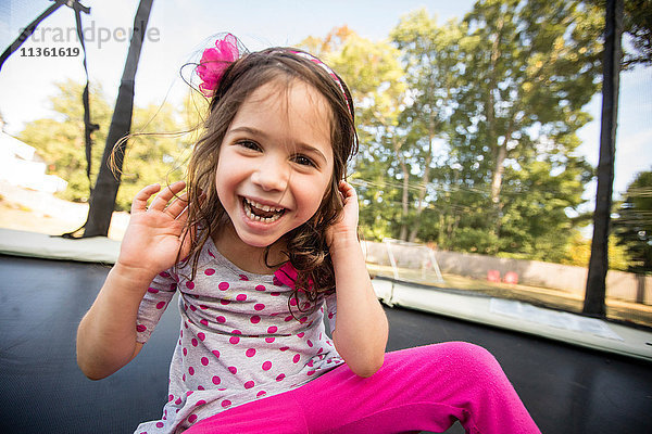 Porträt eines jungen Mädchens,  das auf einem großen Trampolin sitzt und lacht