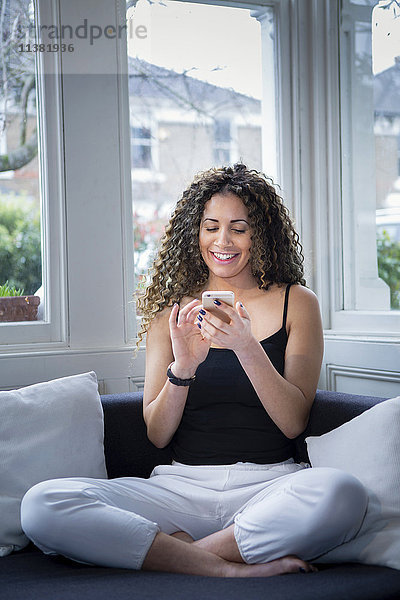 Lächelnde Frau sitzt in der Nähe des Fensters und schreibt eine SMS auf ihrem Handy