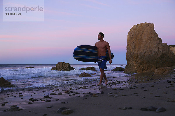 Hispanischer Mann mit Surfbrett am Strand bei Sonnenuntergang