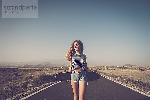 Spanien,  Teneriffa,  lächelnde Frau mit Longboard auf leerer Landstraße stehend