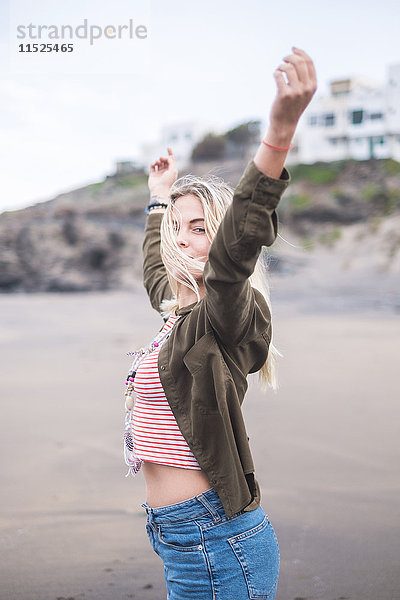 Porträt einer blonden jungen Frau am Strand mit erhobenen Armen