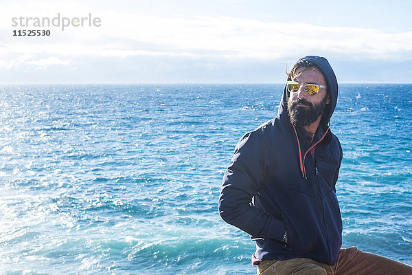 Porträt eines Mannes mit verspiegelter Sonnenbrille und Kapuzenjacke vor dem Meer