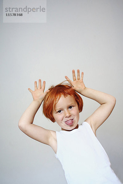 Porträt eines rothaarigen Mädchens im Haus,  das seine Zunge herausstreckt und seine Hände in die Luft legt.