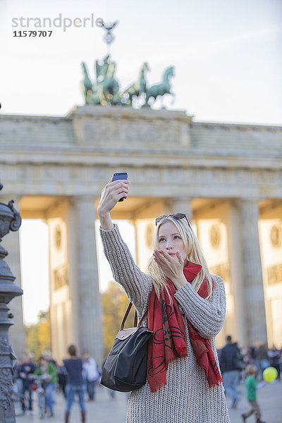 Hübsche blonde Frau beim Selfie und Kuss vor dem Brandenburger Tor in Berlin.