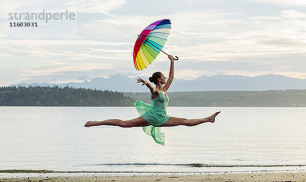 Kaukasische Ballerina springt mit mehrfarbigem Regenschirm am Strand