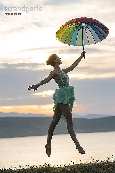 Kaukasische Ballerina springt mit mehrfarbigem Regenschirm am Strand