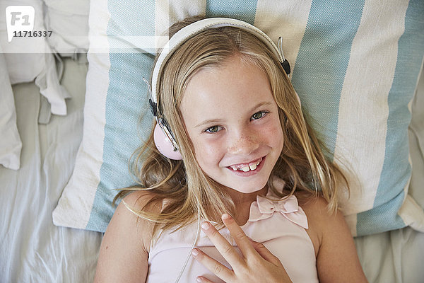 Porträt eines lächelnden kleinen Mädchens,  das zu Hause auf dem Bett liegt und Musik mit Kopfhörern hört.