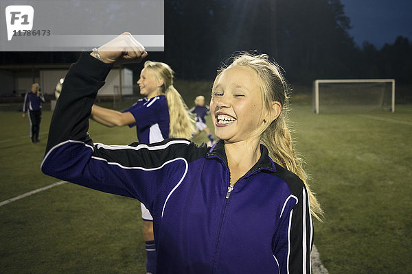 Porträt eines fröhlichen Mädchens,  das seine Muskeln gegen Freunde auf dem Fußballfeld spannt.