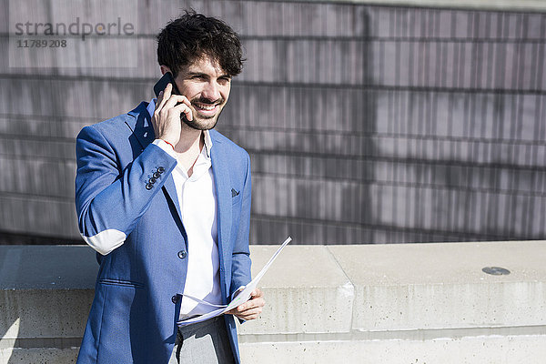Lächelnder junger Geschäftsmann mit Smartphone und Dokumenten im Freien
