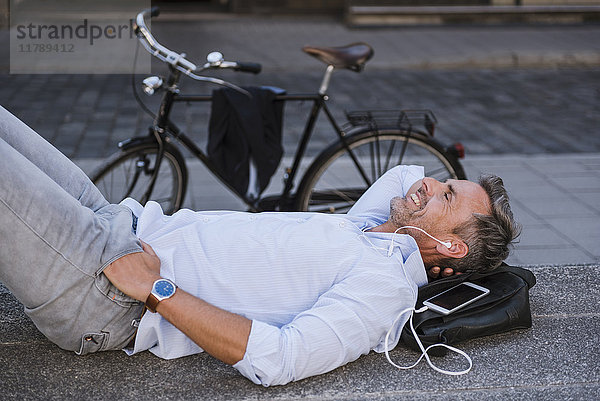 Lächelnder Mann mit geschlossenen Augen auf der Treppe mit Handy und Ohrstöpseln neben dem Fahrrad.