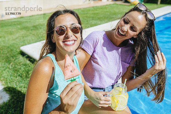 Zwei fröhliche junge Frauen mit Getränk und Wassermelone am Beckenrand