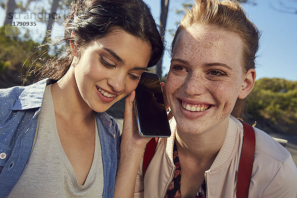 Zwei lächelnde junge Frauen teilen sich ihr Handy im Freien.