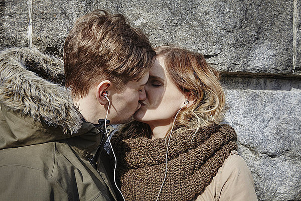 Junges Paar,  das sich die Ohrstöpsel an der Steinmauer küsst