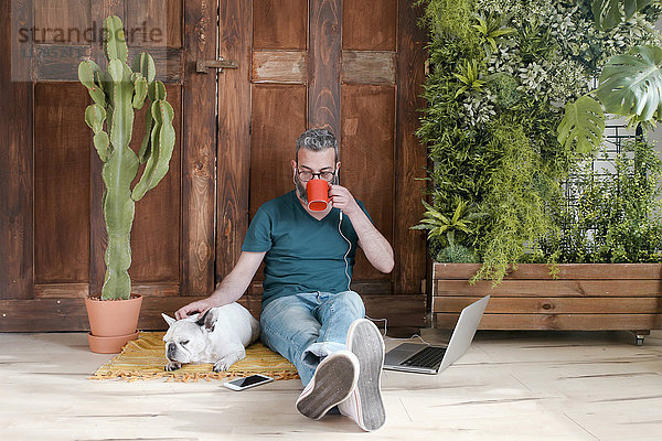 Der bärtige Mann sitzt mit seinem Hund auf dem Boden zu Hause und trinkt Kaffee.
