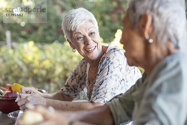 Porträt einer lächelnden Seniorin,  die ihrem Freund auf der Terrasse beim Zubereiten des Mittagessens zuhört.