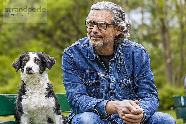 Porträt eines Mannes mit grauem Haar und Bart,  der neben seinem Hund auf einer Bank sitzt.