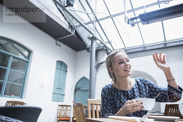 Lächelnde junge Frau winkt und hält eine Kaffeetasse,  während sie im Restaurant sitzt