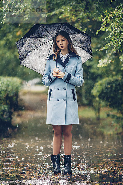 Porträt eines jungen Mädchens mit Regenschirm,  das auf dem Weg zwischen Bäumen steht.