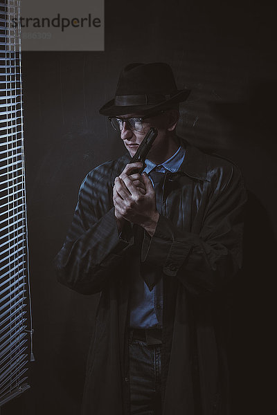 Junger Mann steht mit Pistole am Fenster gegen schwarze Wand