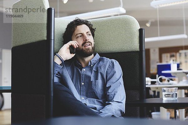 Mittlerer Erwachsener,  professioneller Mann,  der über Smartphone spricht,  während er im Kreativbüro auf dem Stuhl sitzt.