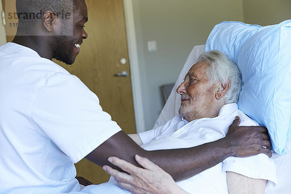 Fröhlicher Krankenpfleger tröstlicher älterer Mann auf dem Bett in der Krankenstation