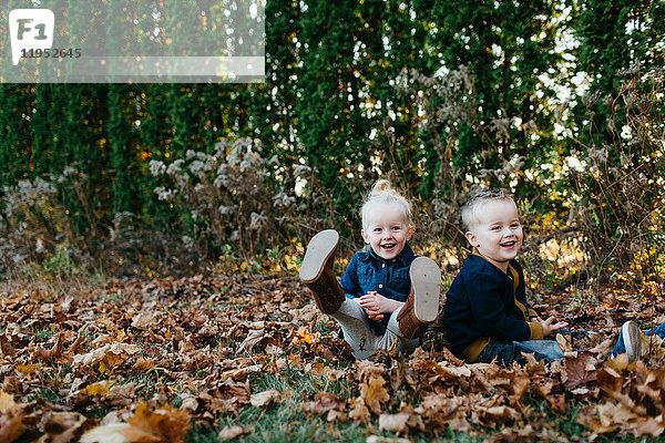 Porträt von männlichen und weiblichen Zwillingskindern,  die zwischen Herbstblättern im Garten sitzen