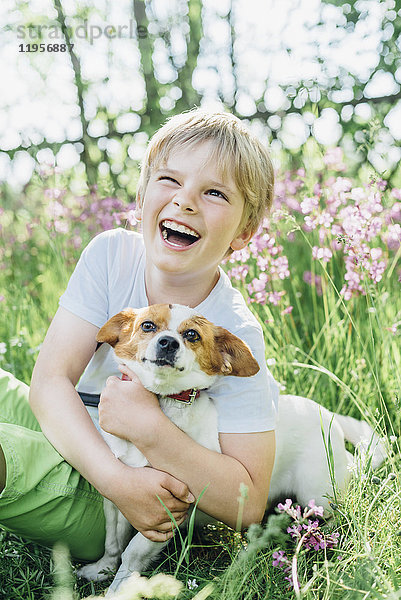 Porträt eines lachenden kleinen Jungen,  der mit seinem Hund auf einer Wiese im Garten sitzt.