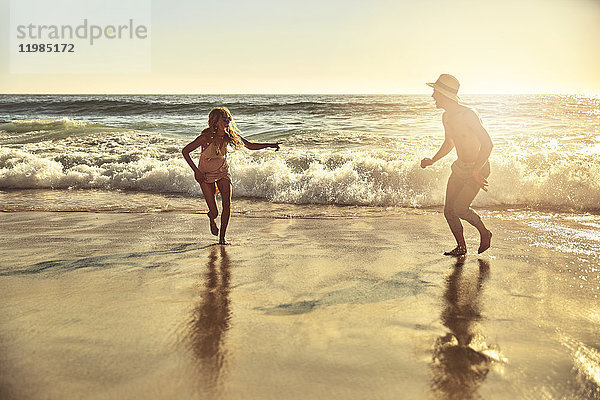 Verspieltes junges Paar,  das im nassen Sand am sonnigen Sommerstrand läuft.